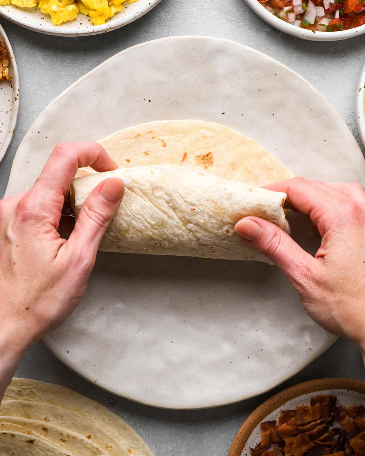 two hands rolling a breakfast burrito on a plate