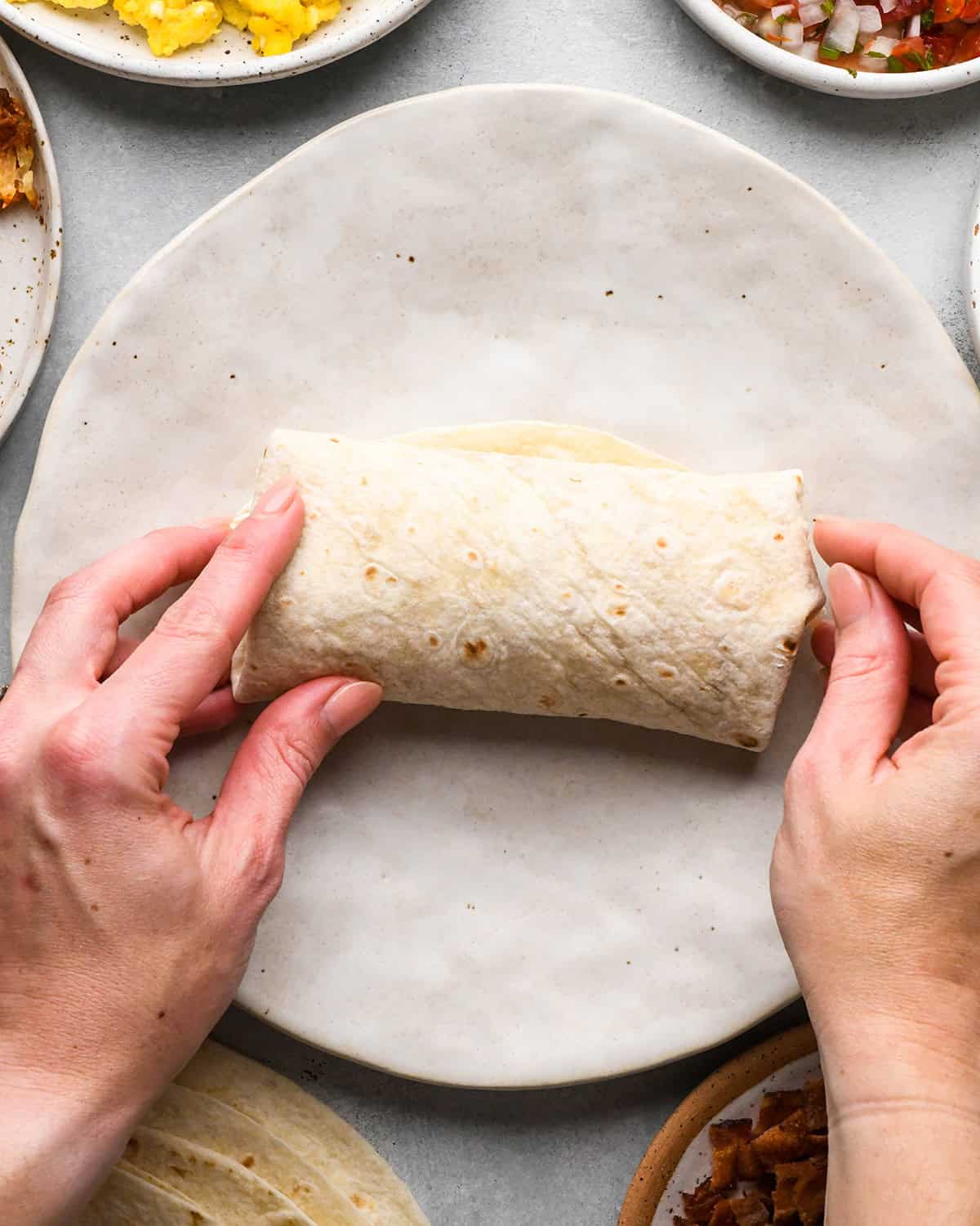 two hands rolling a breakfast burrito on a plate
