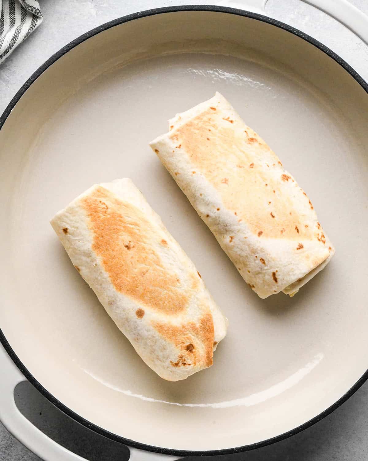 two breakfast burritos cooking on the second side in a skillet