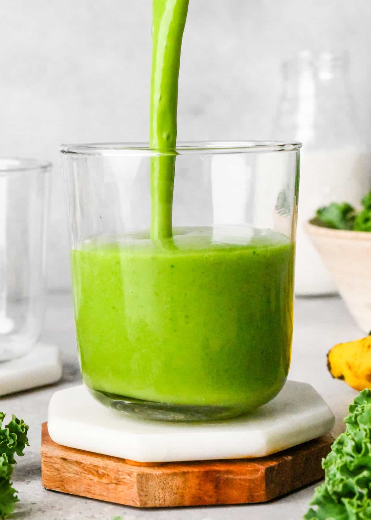 Making kale fruit smoothie: front-view photo of kale fruit smoothie being poured into a clear glass on a white coaster. Kale leaves and bananas surround the glass on a white countertop.