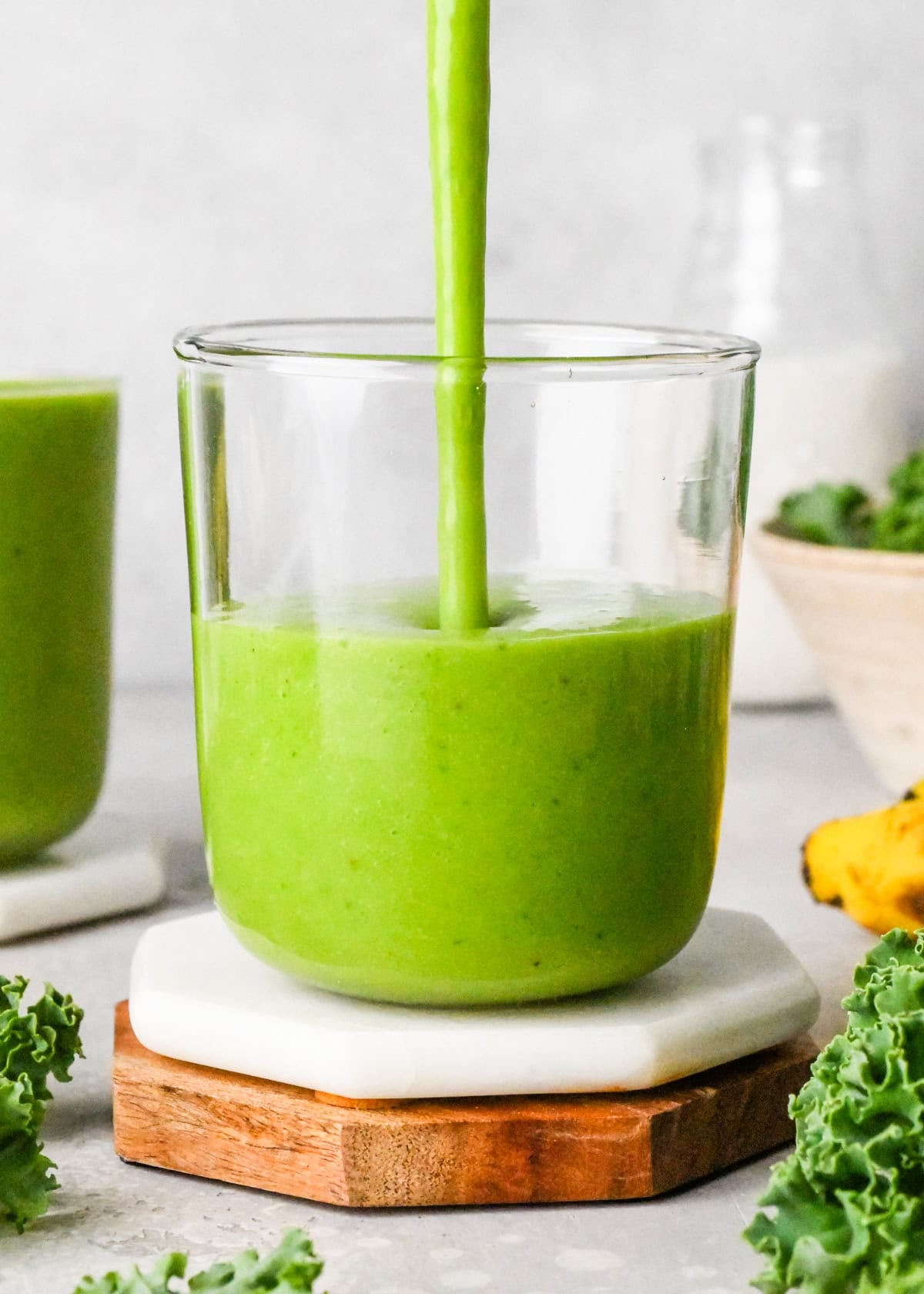 Close-up of a bright green kale fruit smoothie in a glass. The glass is sitting on a white coaster with a straw and there are bananas and fresh kale leaves in the background.