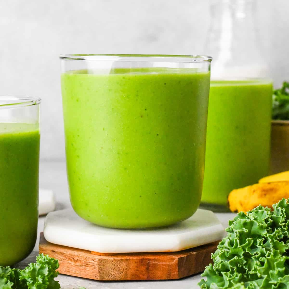 Close-up of a bright green kale fruit smoothie in a glass. The glass is sitting on a white coaster and there are bananas and fresh kale leaves in the background.
