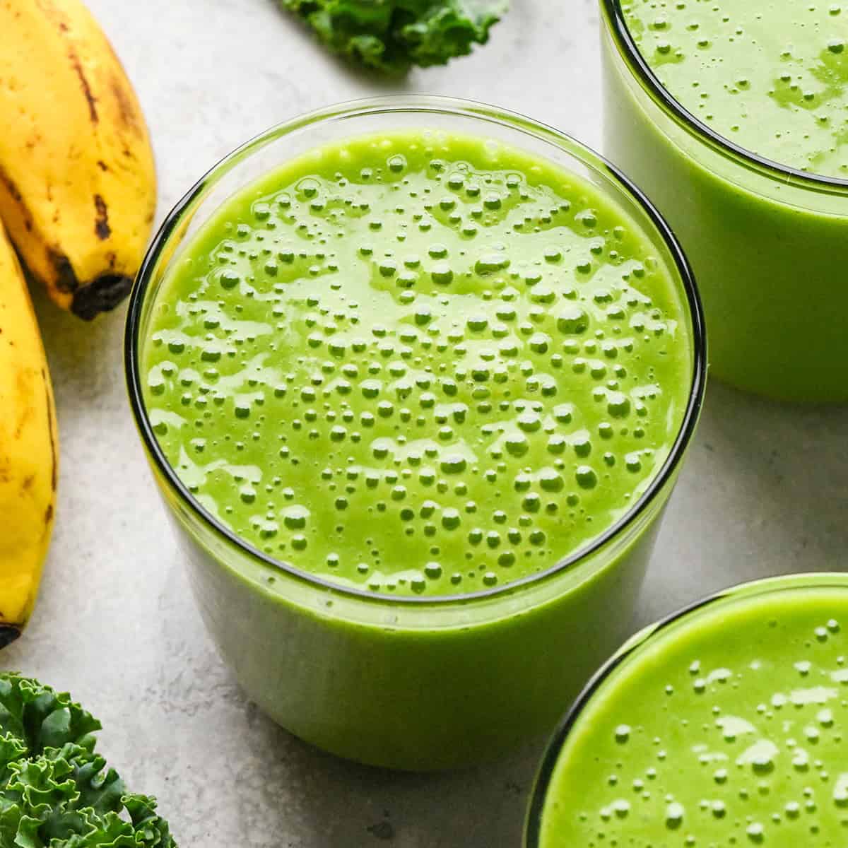 Close-up of a bright green kale fruit smoothie in a glass, with bananas and fresh kale leaves in the background.