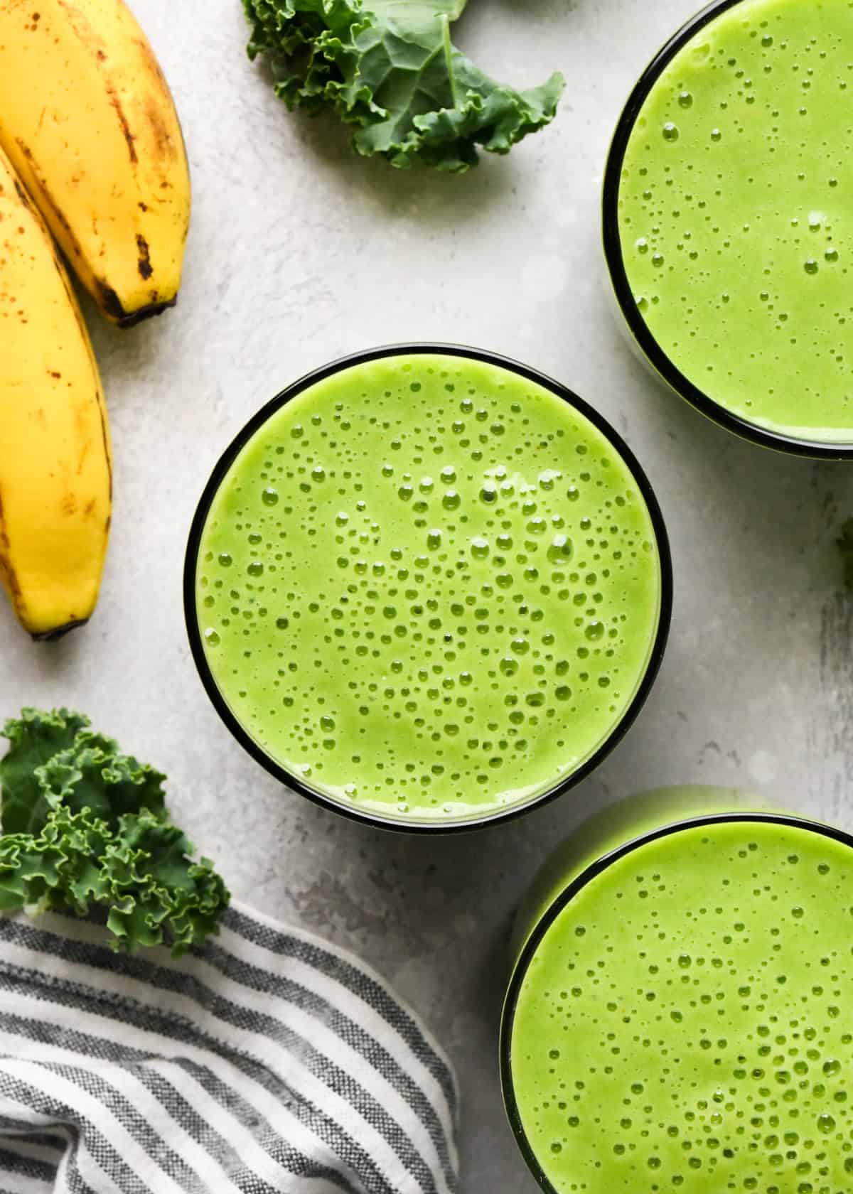 Making kale fruit smoothie: overhead photo of 3 clear glasses filled with bright green kale fruit smoothie and sitting on a countertop.
