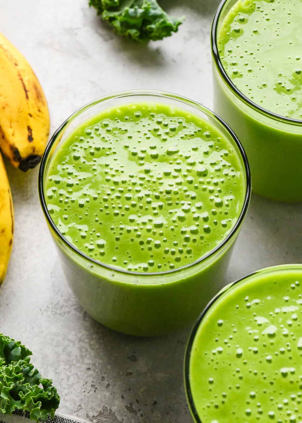 Making kale fruit smoothie: close-up photo of 3 clear glasses filled with bright green kale fruit smoothie and sitting on a countertop.