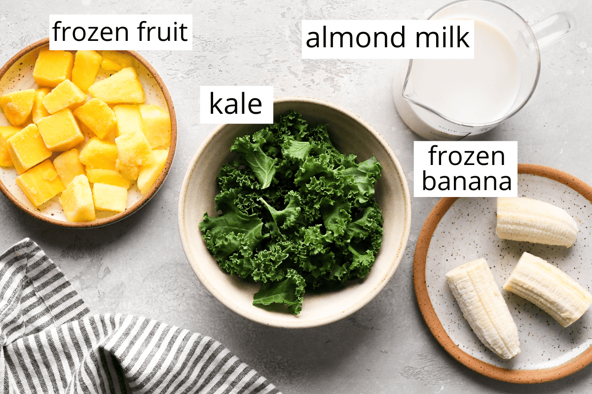 Making kale fruit smoothie: overhead photo of the ingredients for kale fruit smoothie including frozen fruit, kale, frozen banana, and almond milk.