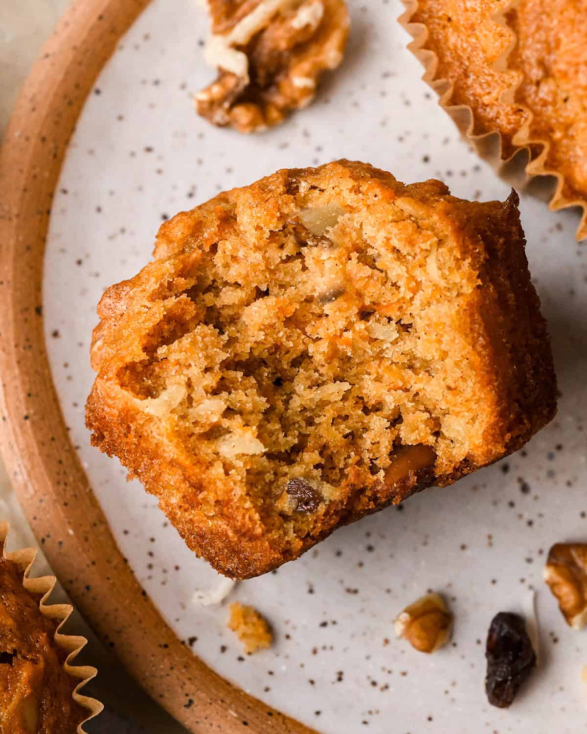 a Morning Glory Muffin on a plate with a bite taken out of it showing the inside texture with grated carrots, apples, raisins, coconut, and chopped nuts surrounded by walnuts, raisins and two other muffins