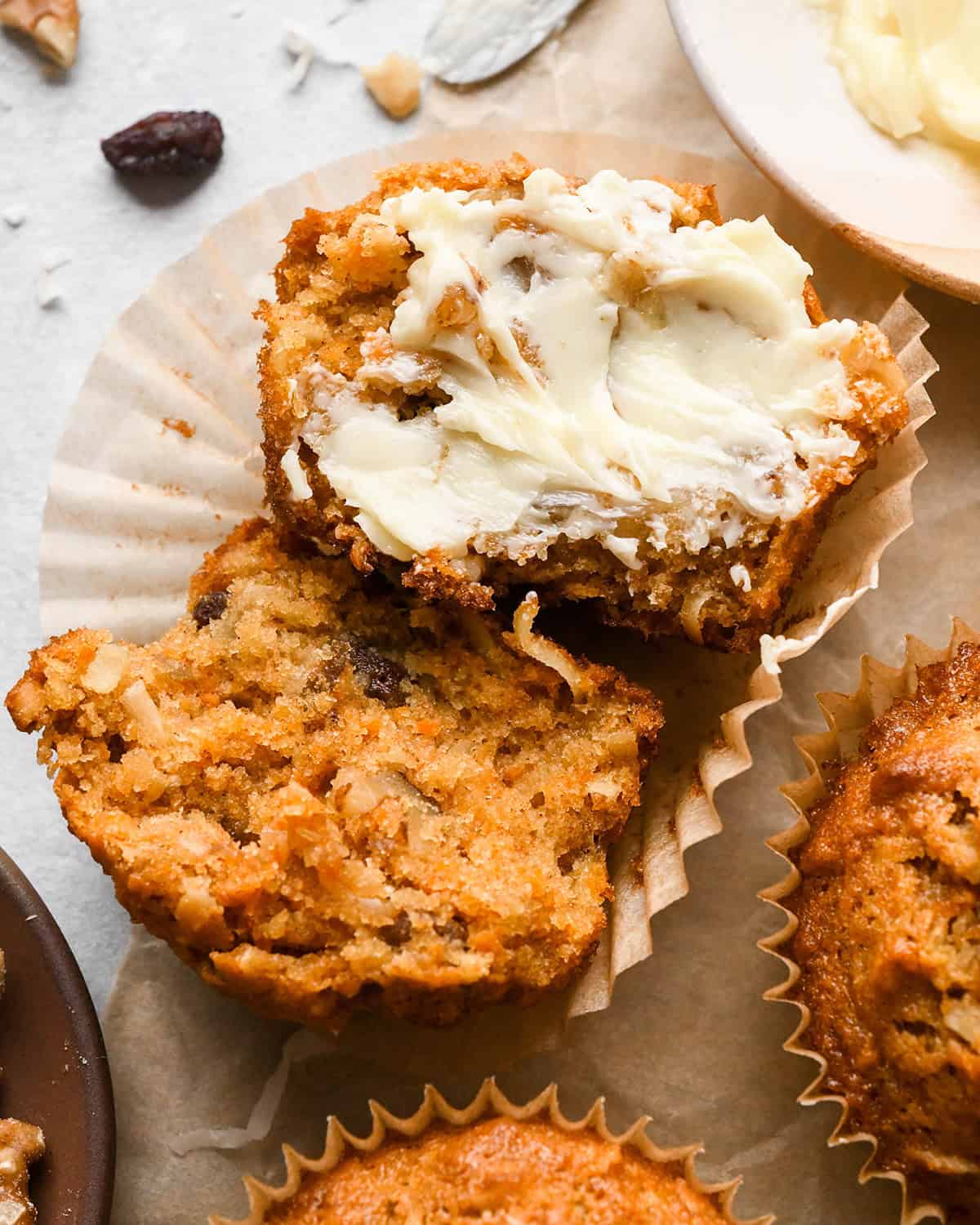 a morning glory muffin cut in half on a muffin wrapper, showing the texture filled with grated carrots, apples, raisins, coconut, and chopped nuts, one half has butter spread on it