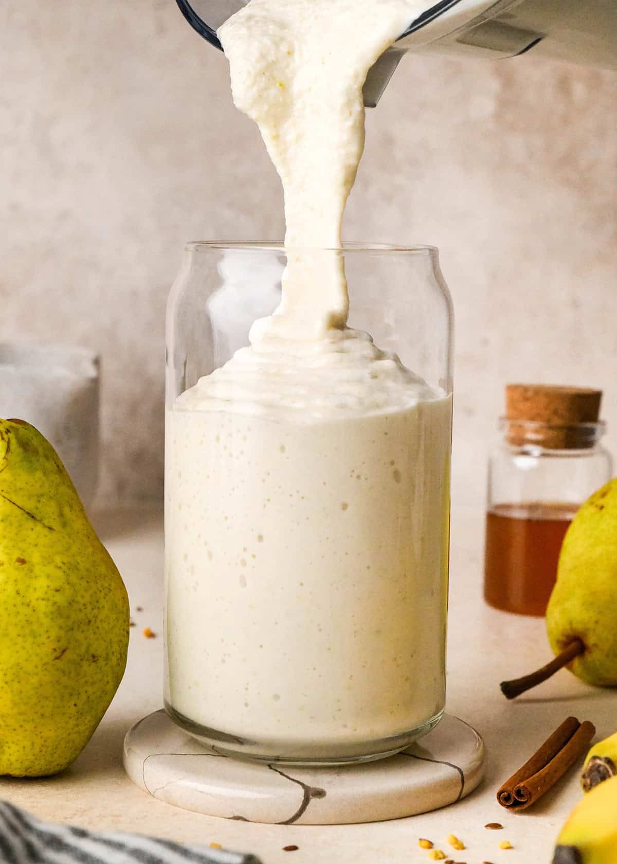front view of Pear Smoothie being poured into a glass surrounded by pears, a cinnamon stick  and honey