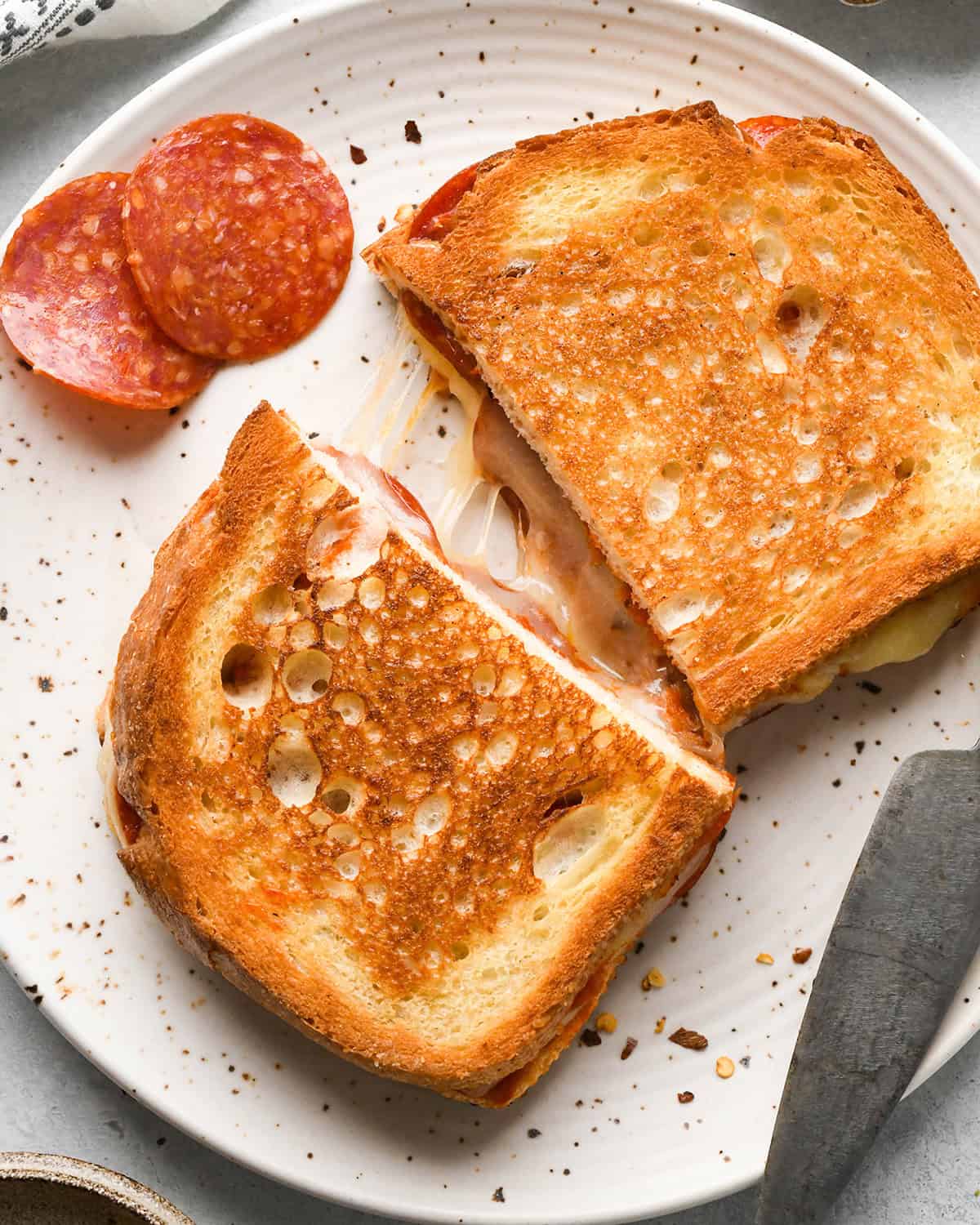 a pizza grilled cheese cut in half on a white plate with melted cheese between the two halves and pepperoni on the plate