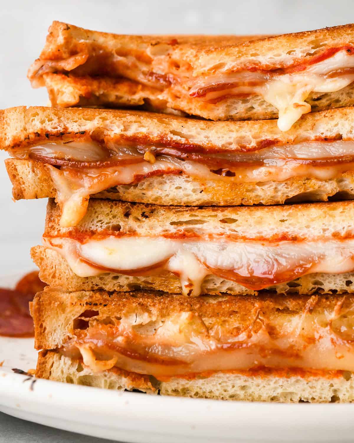 Stack of 4 halves of pizza grilled cheese sandwiches filled with melted mozzarella cheese, pepperoni, and pizza sauce, between slices of toasted bread on a white plate. The top one has a bite taken out of it