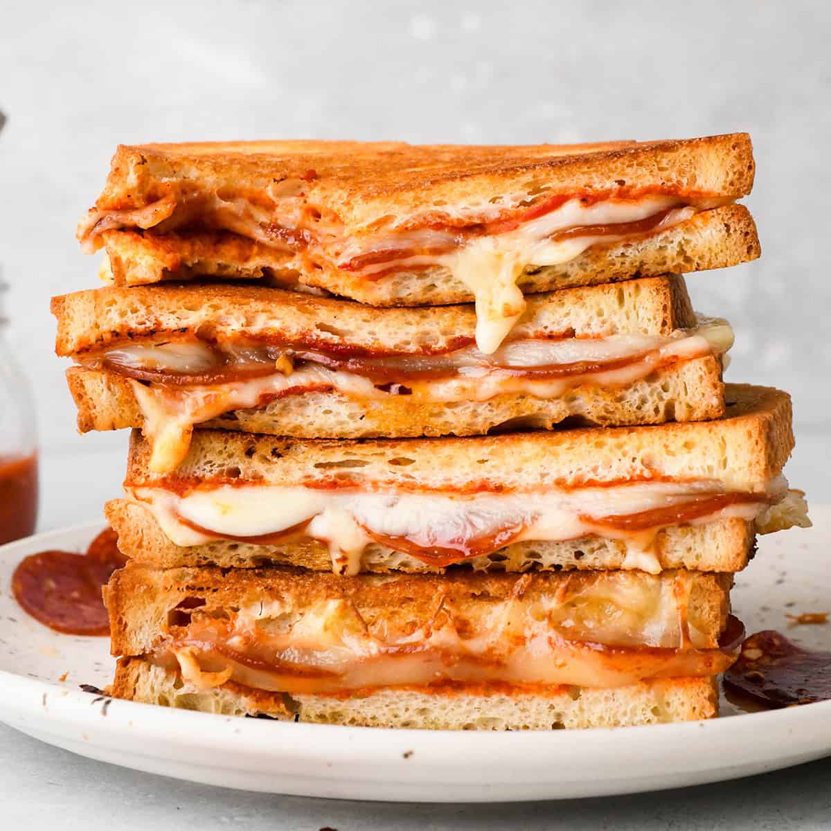 Stack of 4 halves of pizza grilled cheese sandwiches filled with melted mozzarella cheese, pepperoni, and pizza sauce, between slices of toasted bread on a white plate
