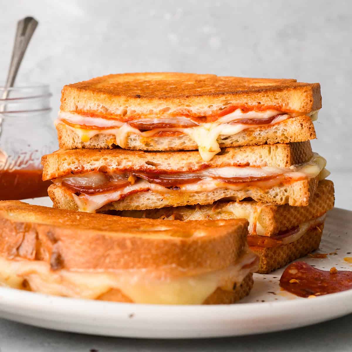 Stack of 3 halves of pizza grilled cheese sandwiches filled with melted mozzarella cheese, pepperoni, and pizza sauce, between slices of toasted bread on a white plate with pepperoni and another pizza grilled cheese half on the plate