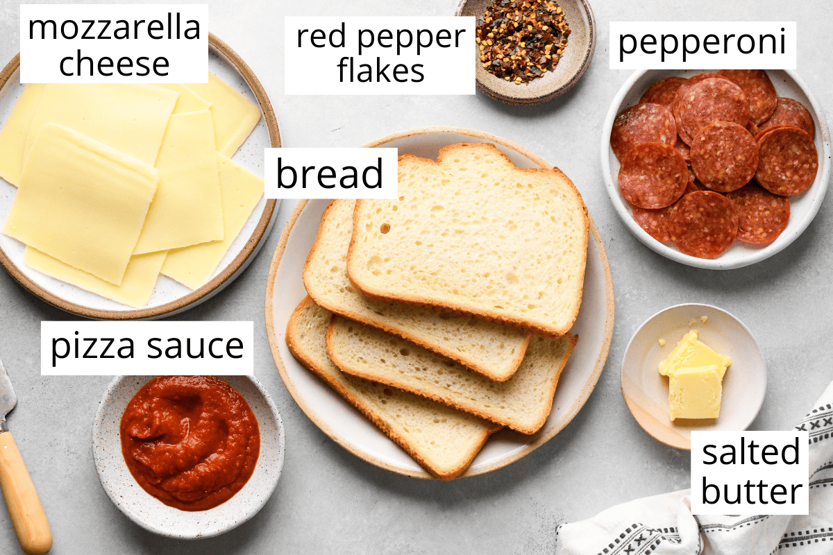 overhead photo of the labeled ingredients in this Pizza Grilled Cheese recipe which include mozzarella cheese, bread, red pepper flakes, pepperoni, butter and pizza sauce