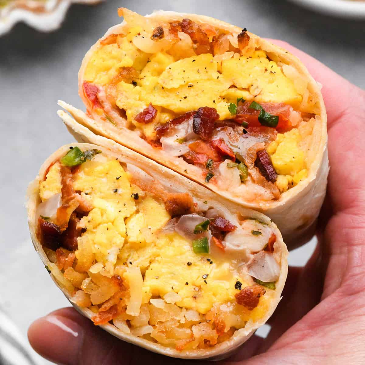 a hand holding a Breakfast Burrito cut in half so you can see it's filled with scrambled eggs, hash browns, bacon, and cheese