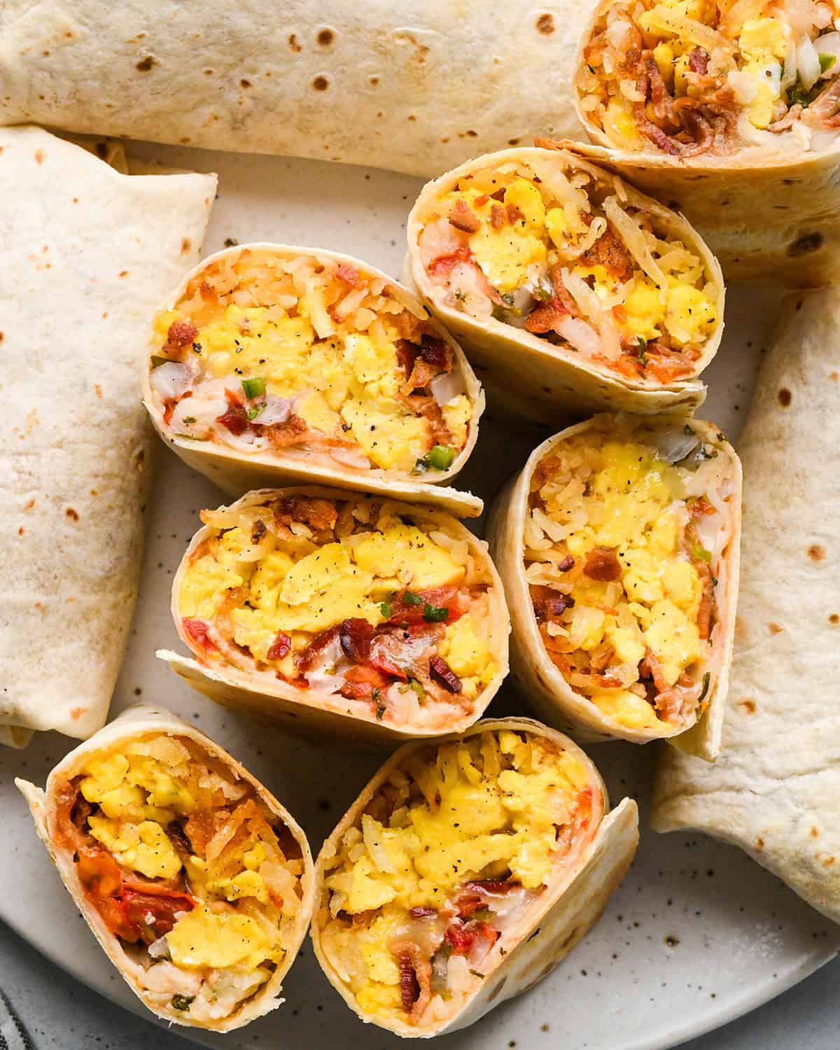 7 breakfast burritos, 4 cut in half showing they're filled with scrambled eggs, hash browns, bacon, and cheese
