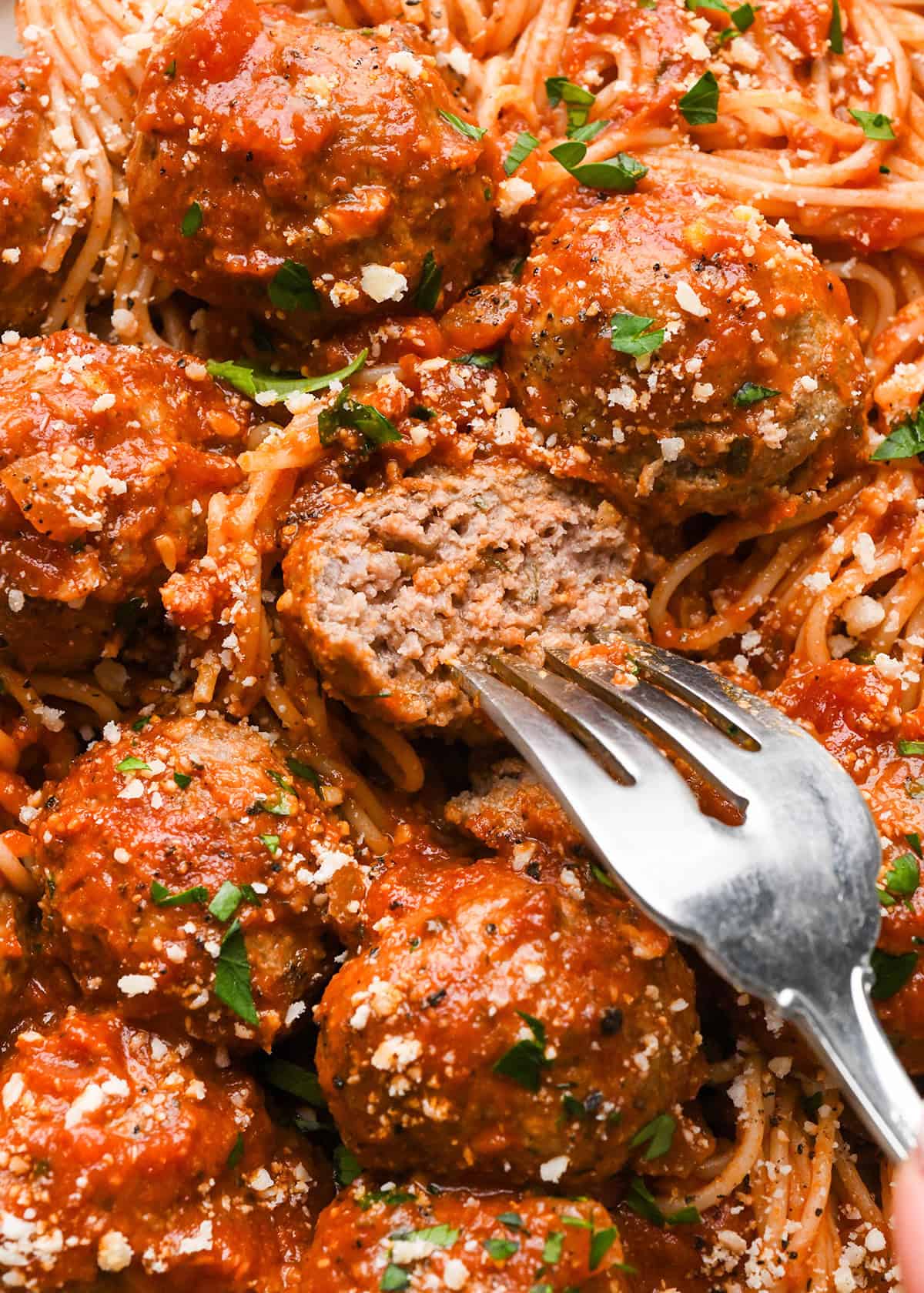 slow cooker meatballs in marinara sauce, served over spaghetti and sprinkled with grated parmesan cheese and fresh parsley. One meatball is cut in half to show the moist interior and a fork is in that meatball