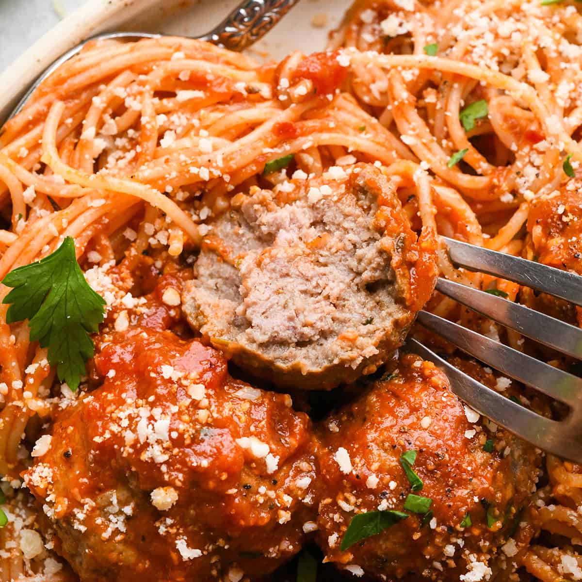 slow cooker meatballs in marinara sauce, served over spaghetti and sprinkled with grated parmesan cheese and fresh parsley. One meatball is cut in half to show the moist interior and a fork is in that meatball