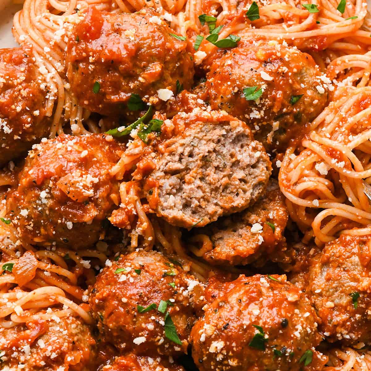 Close-up of slow cooker meatballs in marinara sauce, served over spaghetti and sprinkled with grated parmesan cheese and fresh parsley. One meatball is cut open to show the juicy interior.