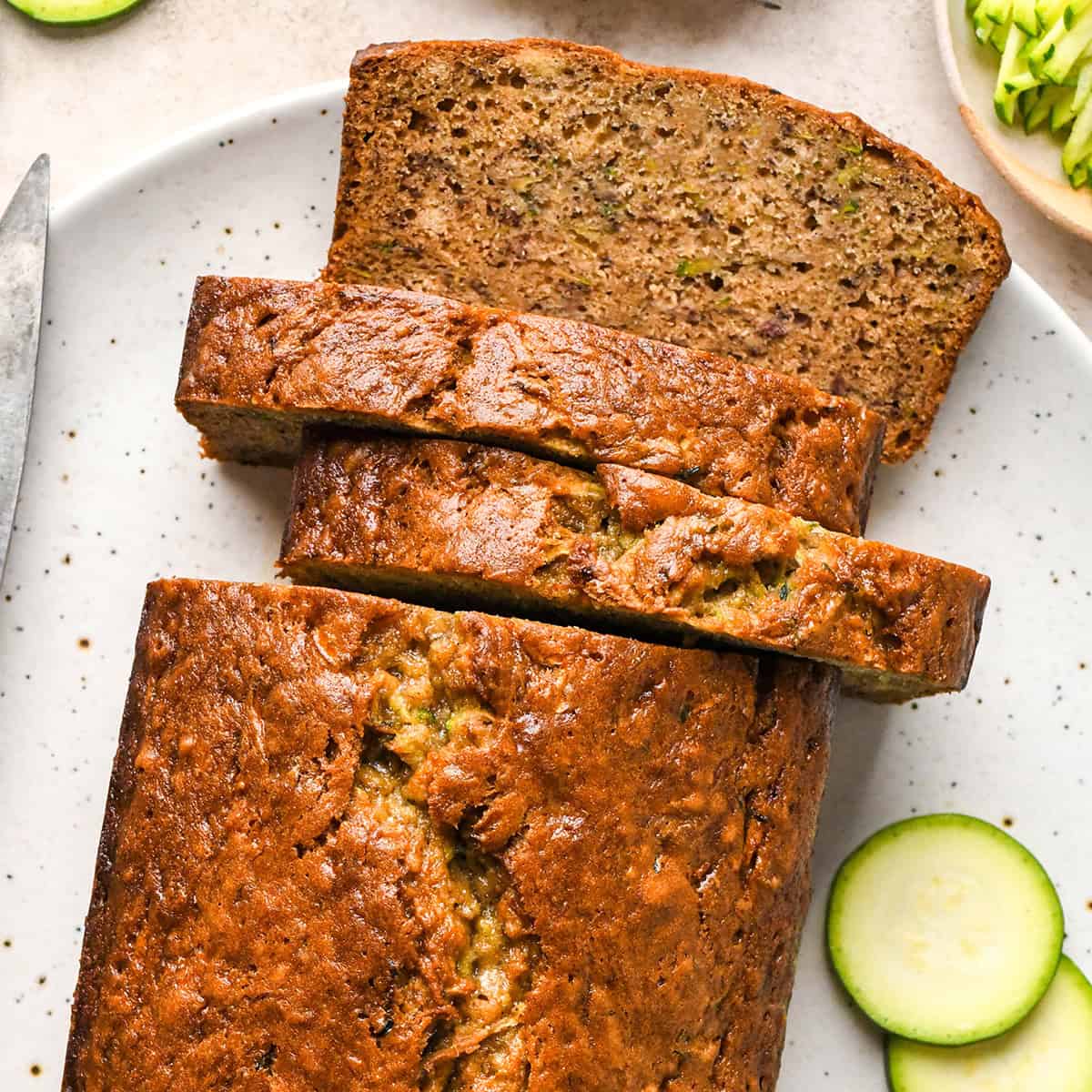 a loaf of Zucchini Banana Bread with 3 slices cut out of it on a white plate with slices of zucchini.