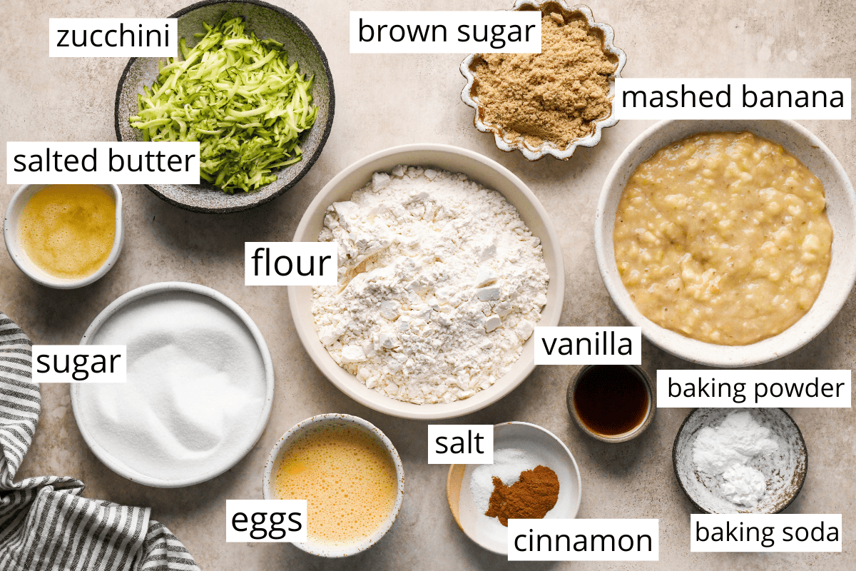 overhead photo of the labeled ingredients in this Zucchini Banana Bread recipe like zucchini, mashed banana, flour, brown sugar, butter, sugar, and mroe