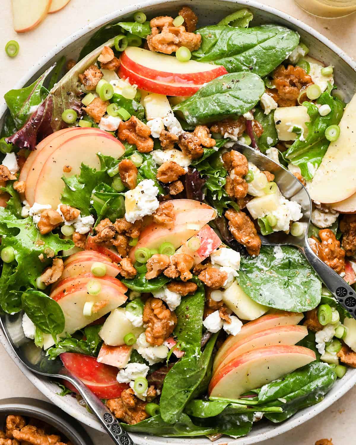 apple salad with mixed greens, thinly sliced apples, crumbled goat cheese, candied walnuts, chopped green onions, and a vinaigrette dressing, served in a white bowl with two serving spoons