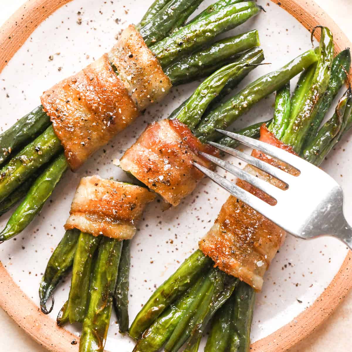 3 bundles of bacon wrapped green beans sprinkled with salt and black pepper on a white plate. the middle one is cut in half and a fork is sticking into it 