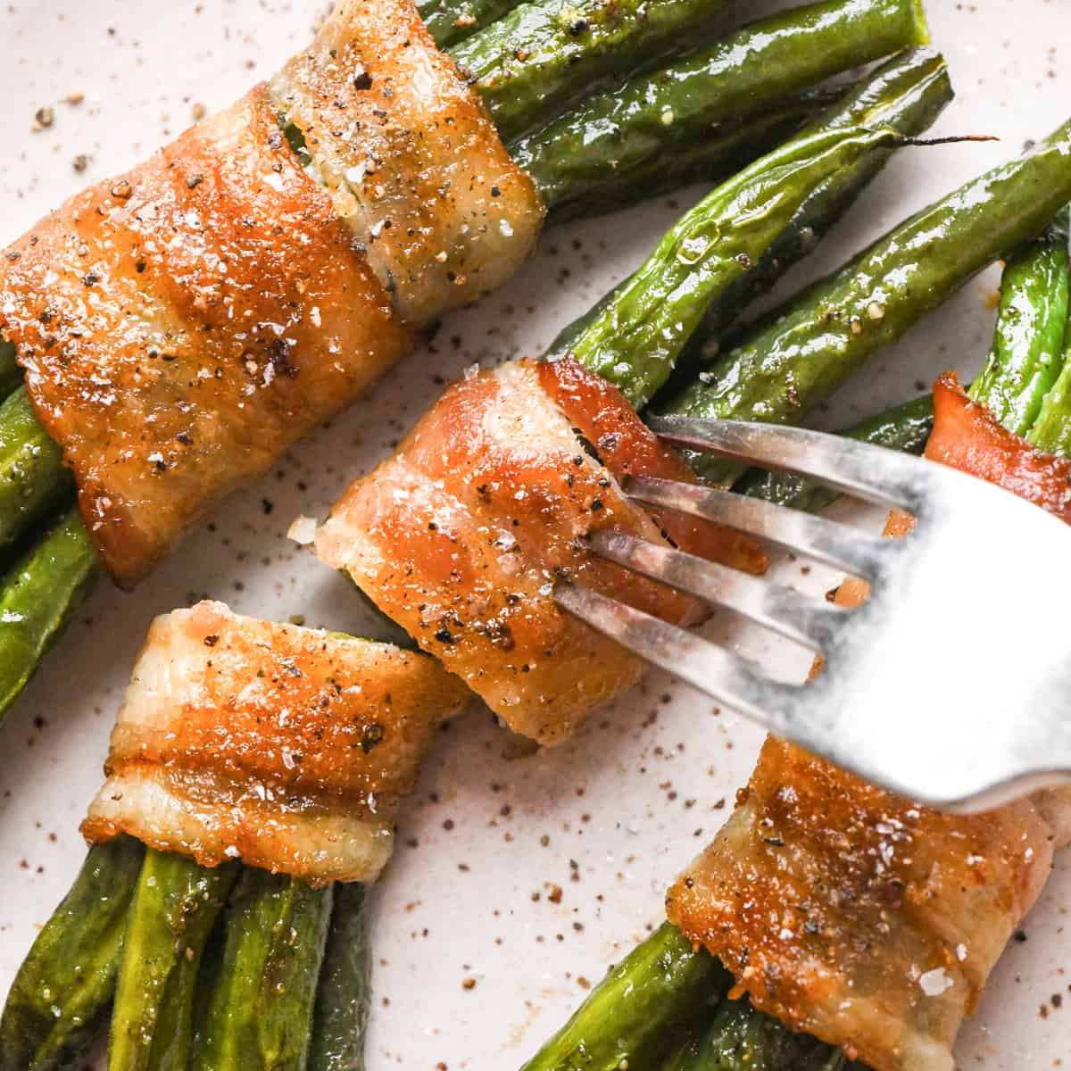 3 bundles of bacon wrapped green beans sprinkled with salt and black pepper on a white plate. the middle one is cut in half and a fork is sticking into it 
