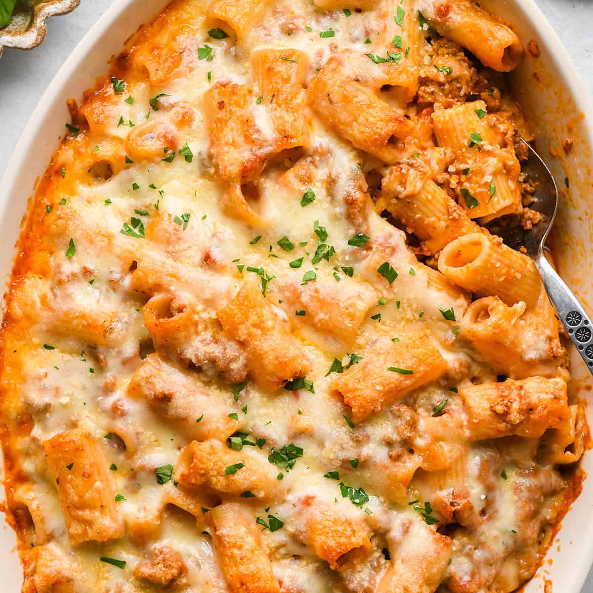 Baked Mostaccioli in a white baking dish with a spoon, garnished with chopped parsley