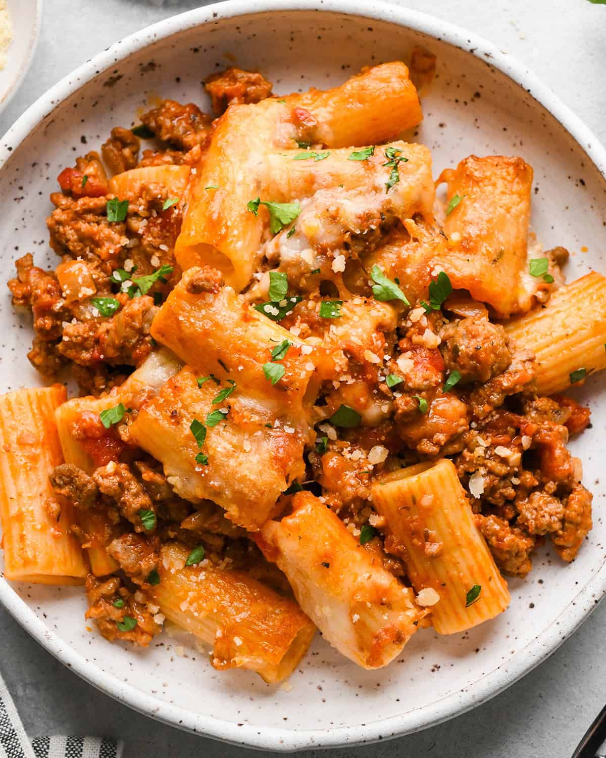 a serving of Baked Mostaccioli on a white plate garnished with chopped parsley and parmesan cheese