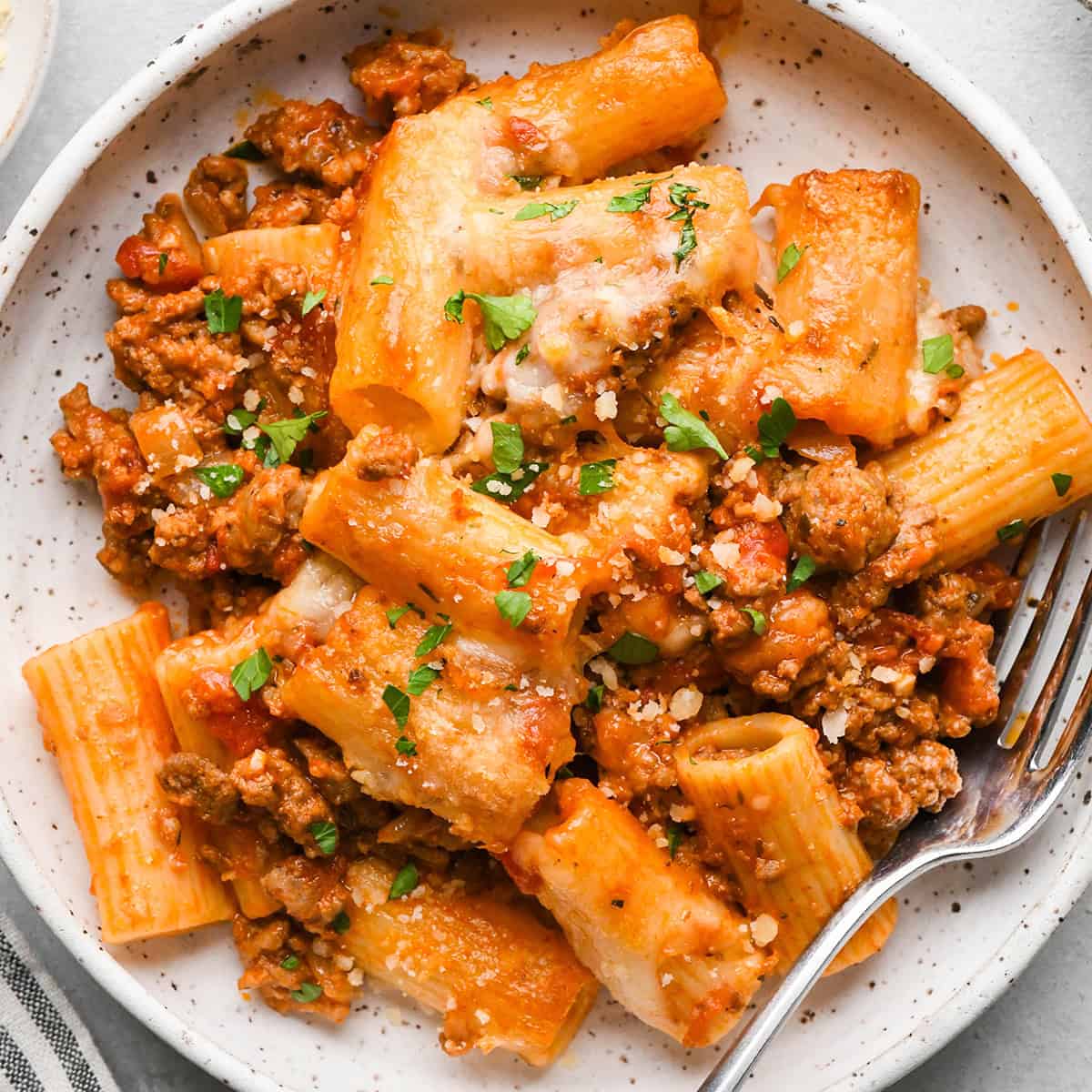 Baked Mostaccioli garnished with parsley and parmesan cheese on a white plate with a fork. 