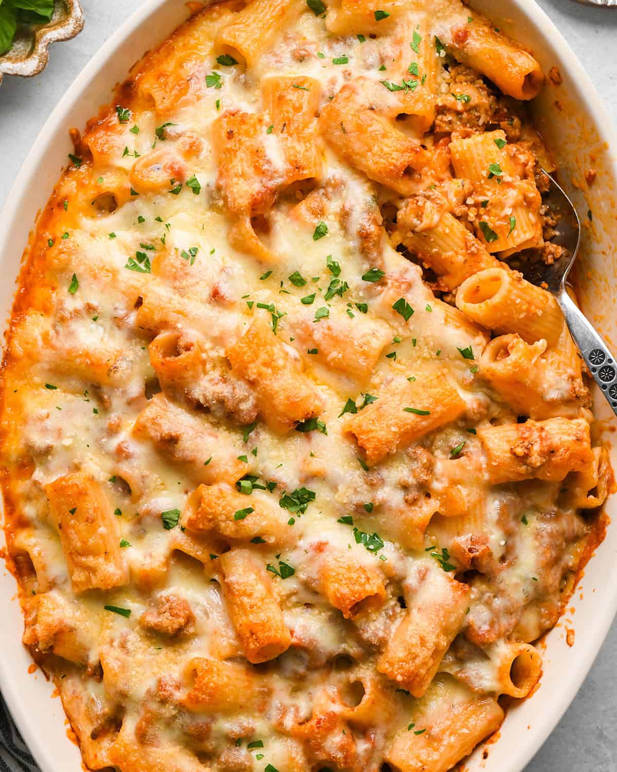 Baked Mostaccioli in a white baking dish with a spoon, garnished with chopped parsley