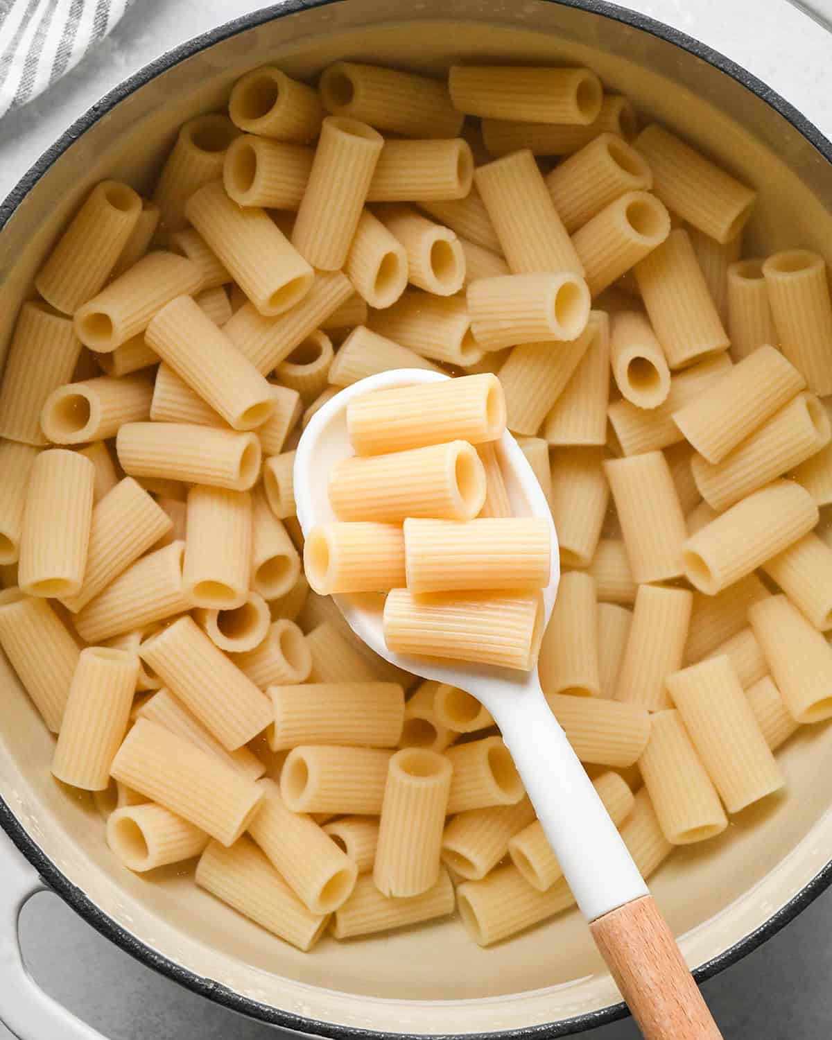 a spoon holding some Mostaccioli noodles cooked over more noodles in a pot of water. 