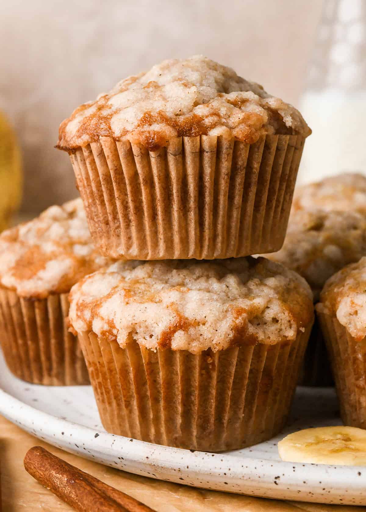 5 banana crumb muffins with a streusel topping, stacked on a white plate with bananas and cinnamon sticks in the background