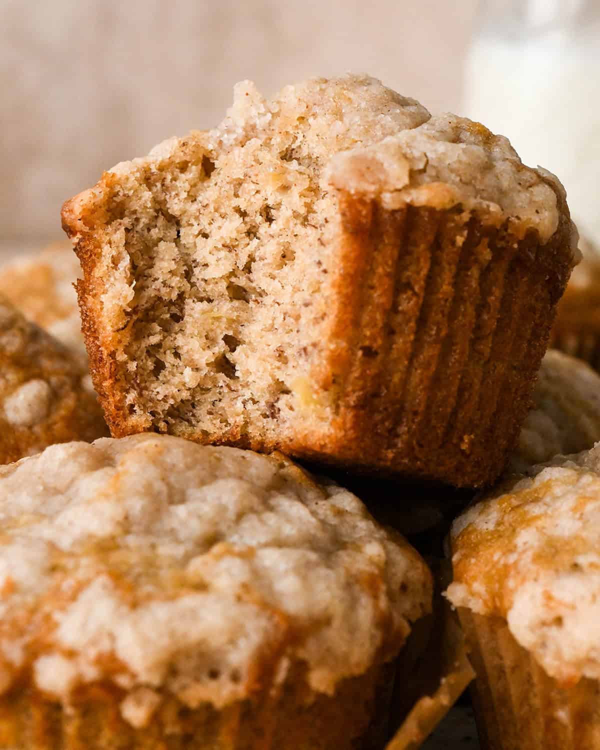 Close-up of a moist banana crumb muffin with a crumb topping with a bite taken out, showing the moist texture. Surrounded by whole muffins.