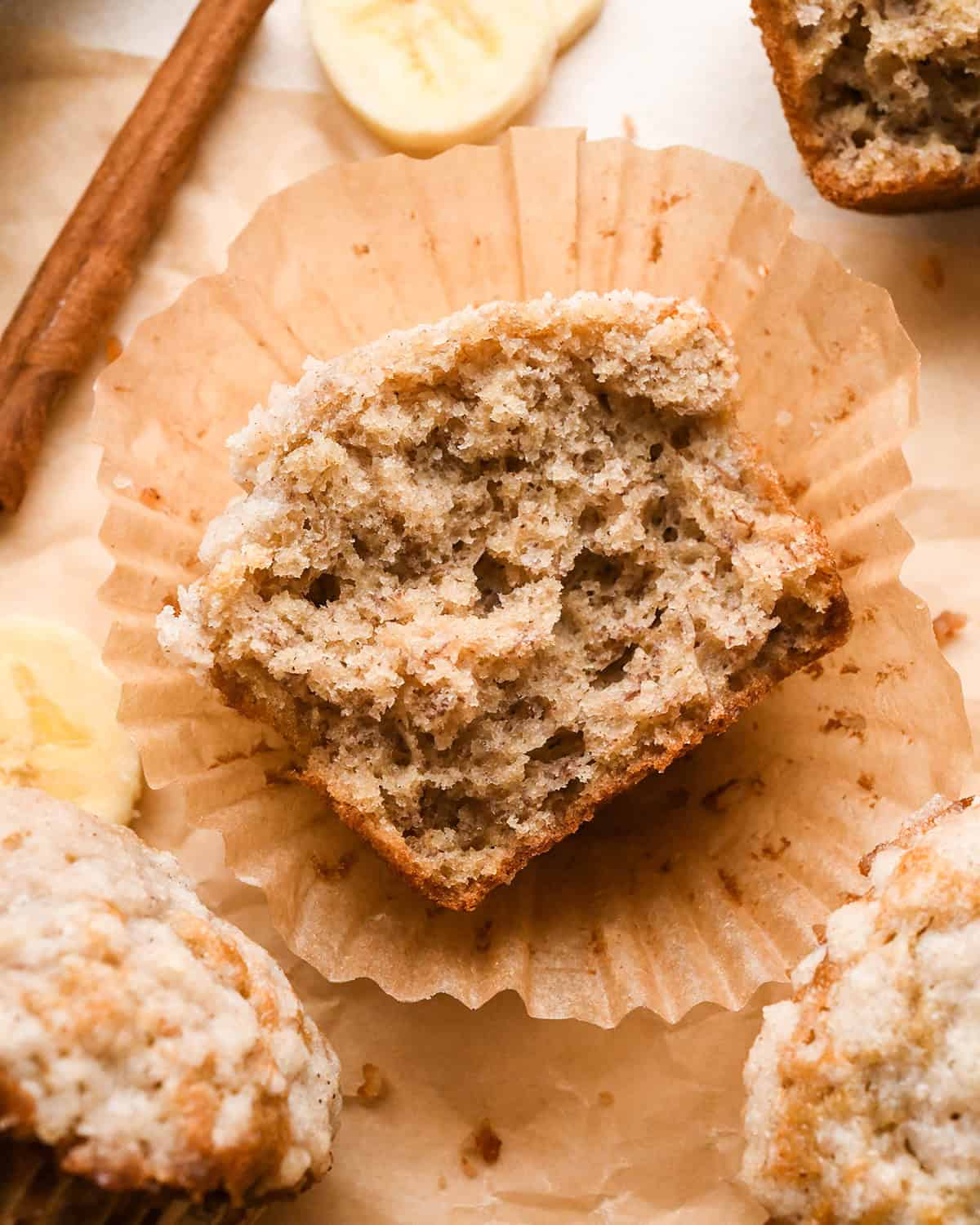 half a banana muffin on a cupcake liner laying down so the moist, fluffy texture is visible surrounded by other muffins, a cinnamon stick and bananas
