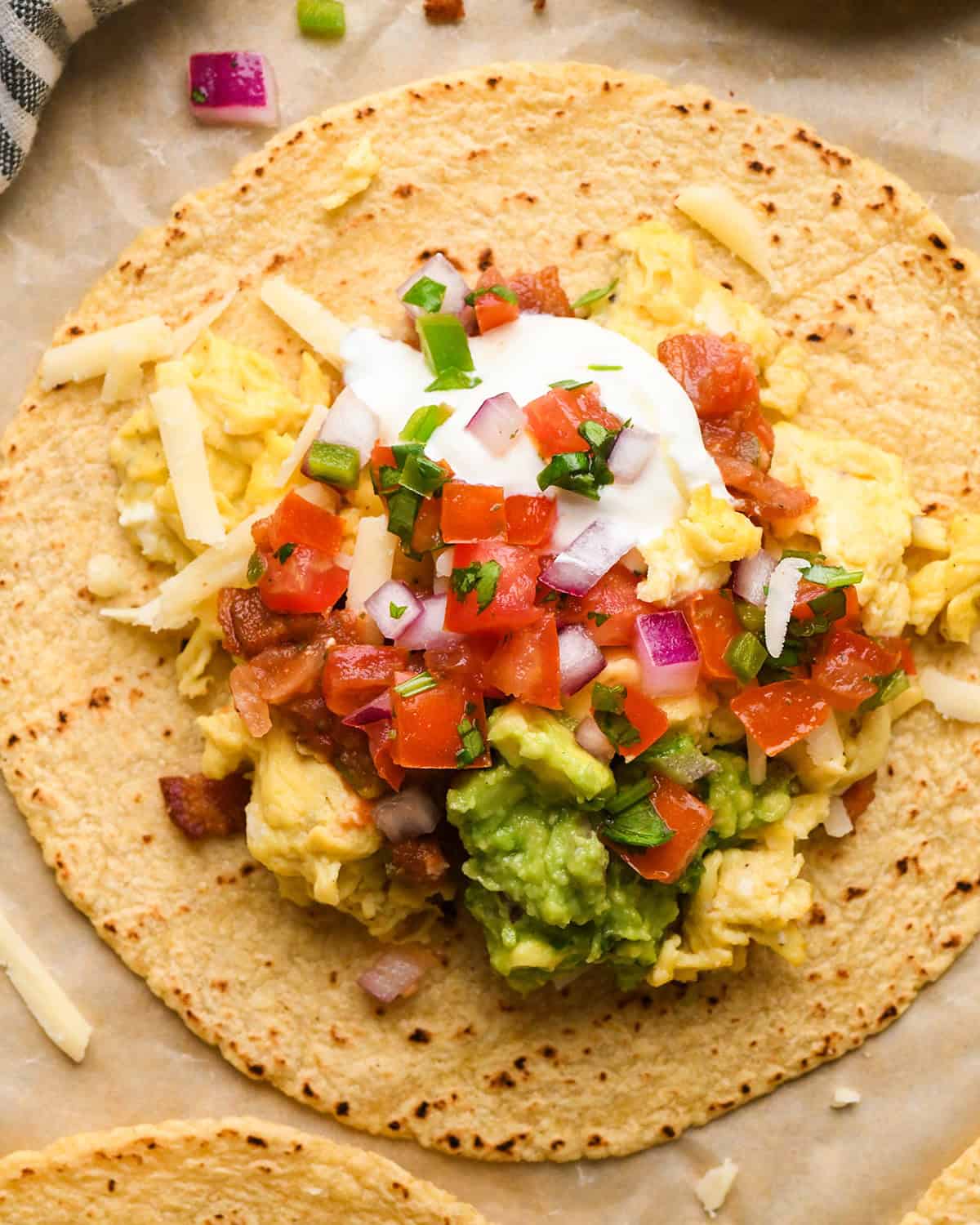 up close photo of a breakfast taco on a corn tortilla filled with scrambled eggs, shredded cheese, bacon, guacamole, pico de gallo, sour cream, and chopped red onions on parchment paper