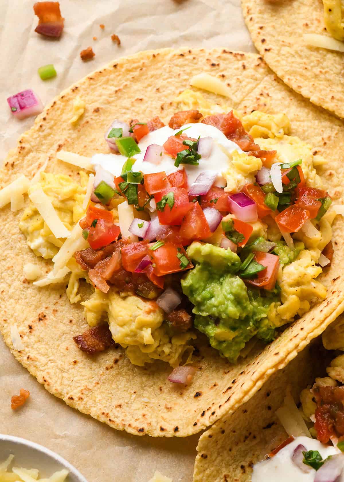 close up photo of a breakfast taco on corn tortillas filled with scrambled eggs, shredded cheese, bacon, guacamole, pico de gallo, sour cream, and chopped red onions