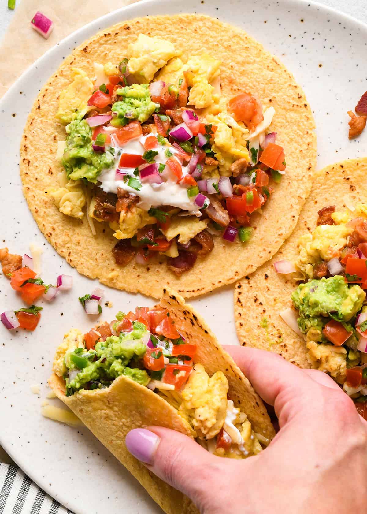 3 breakfast tacos on corn tortillas filled with scrambled eggs, shredded cheese, bacon, guacamole, pico de gallo, sour cream, and chopped red onions on a white plate, a hand is holding one taco and it has a bite taken out of it