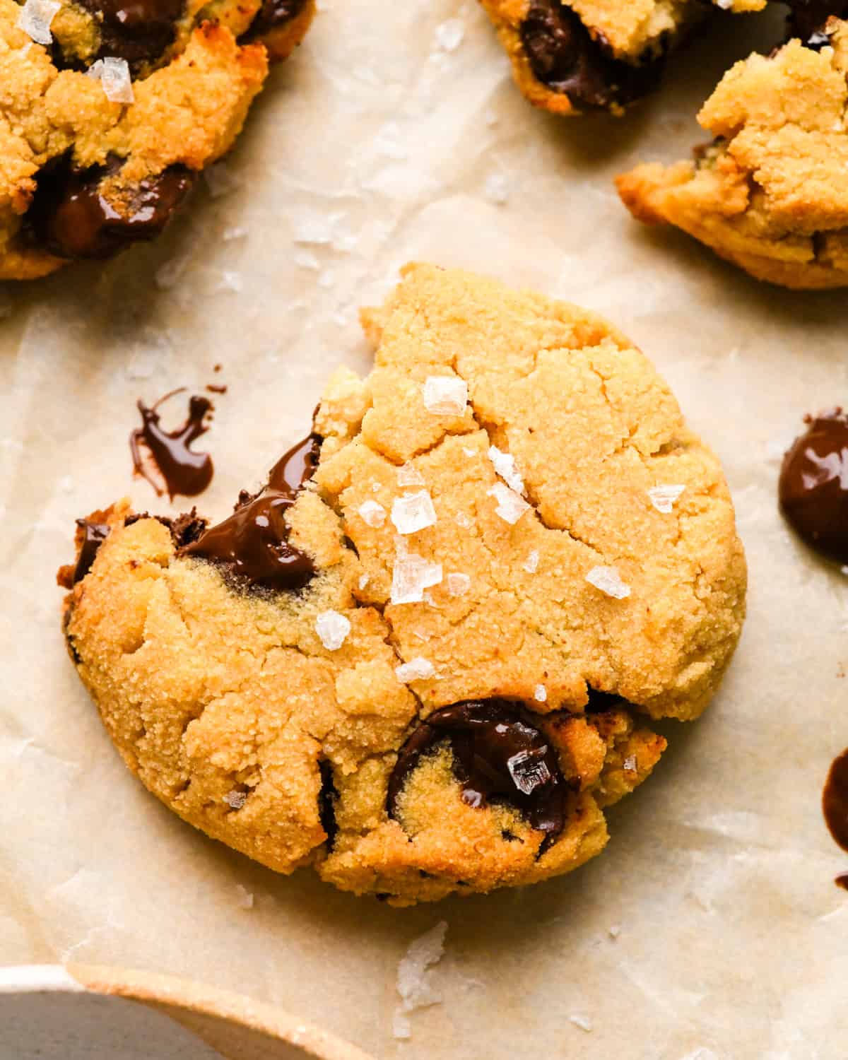 close up photo of a chocolate chip coconut flour cookie with a bite taken out of it topped with flaky sea salt on parchment paper with two other coconut flour cookies and chocolate chips nearby