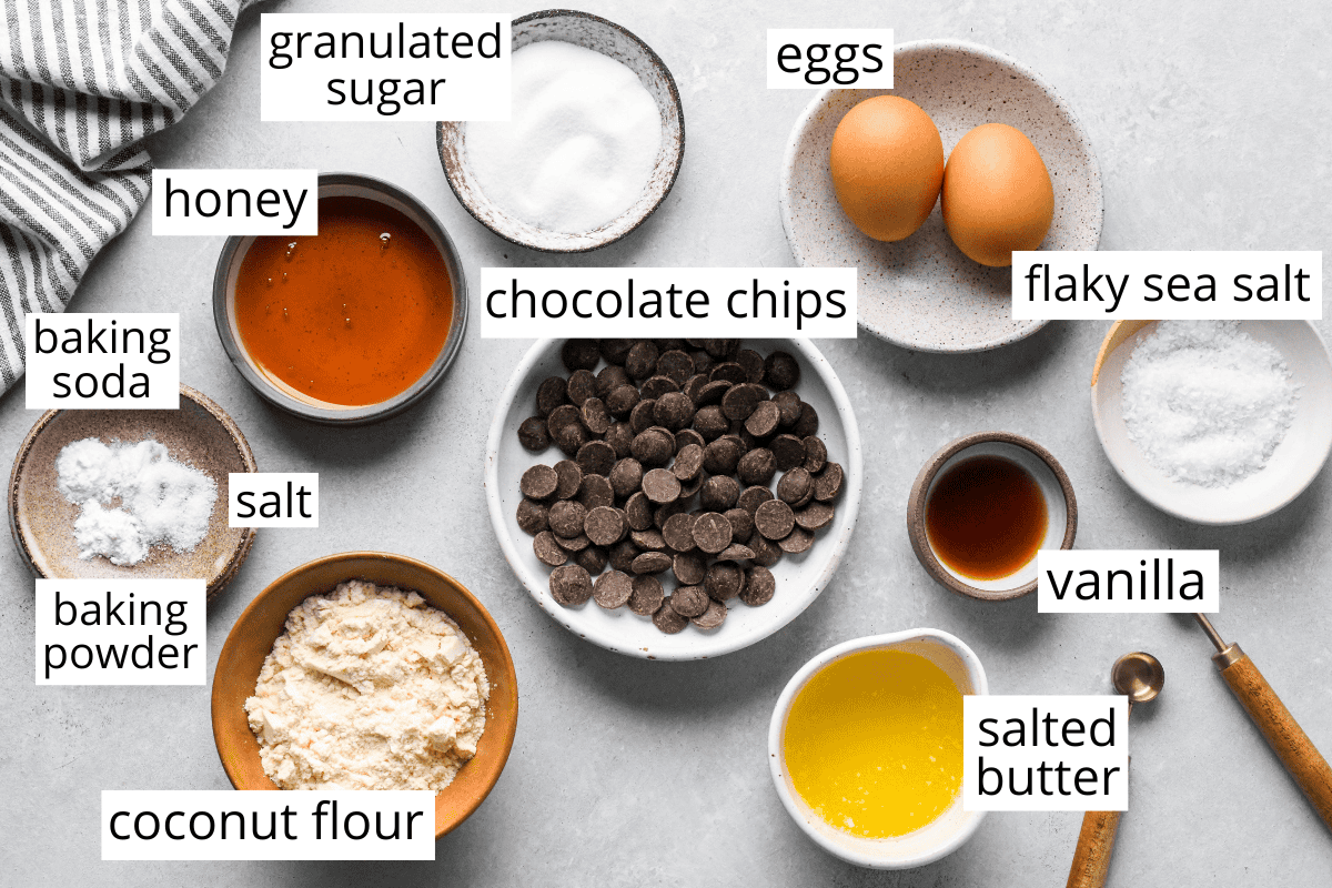 overhead photo of the labeled ingredients in bowls in this Coconut Flour Cookies recipe like chocolate chips, butter, coconut flour, eggs, sugar, honey and flaky sea salt