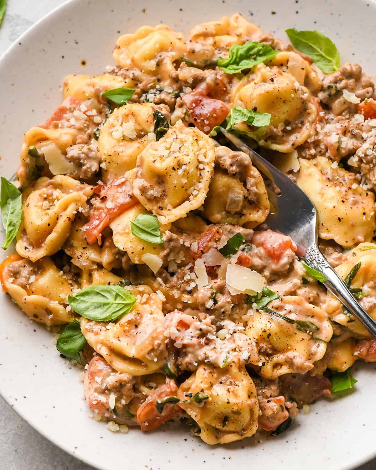 ground beef tortellini with diced tomatoes, fresh basil, and grated parmesan cheese,  in a white bowl with a silver fork. 