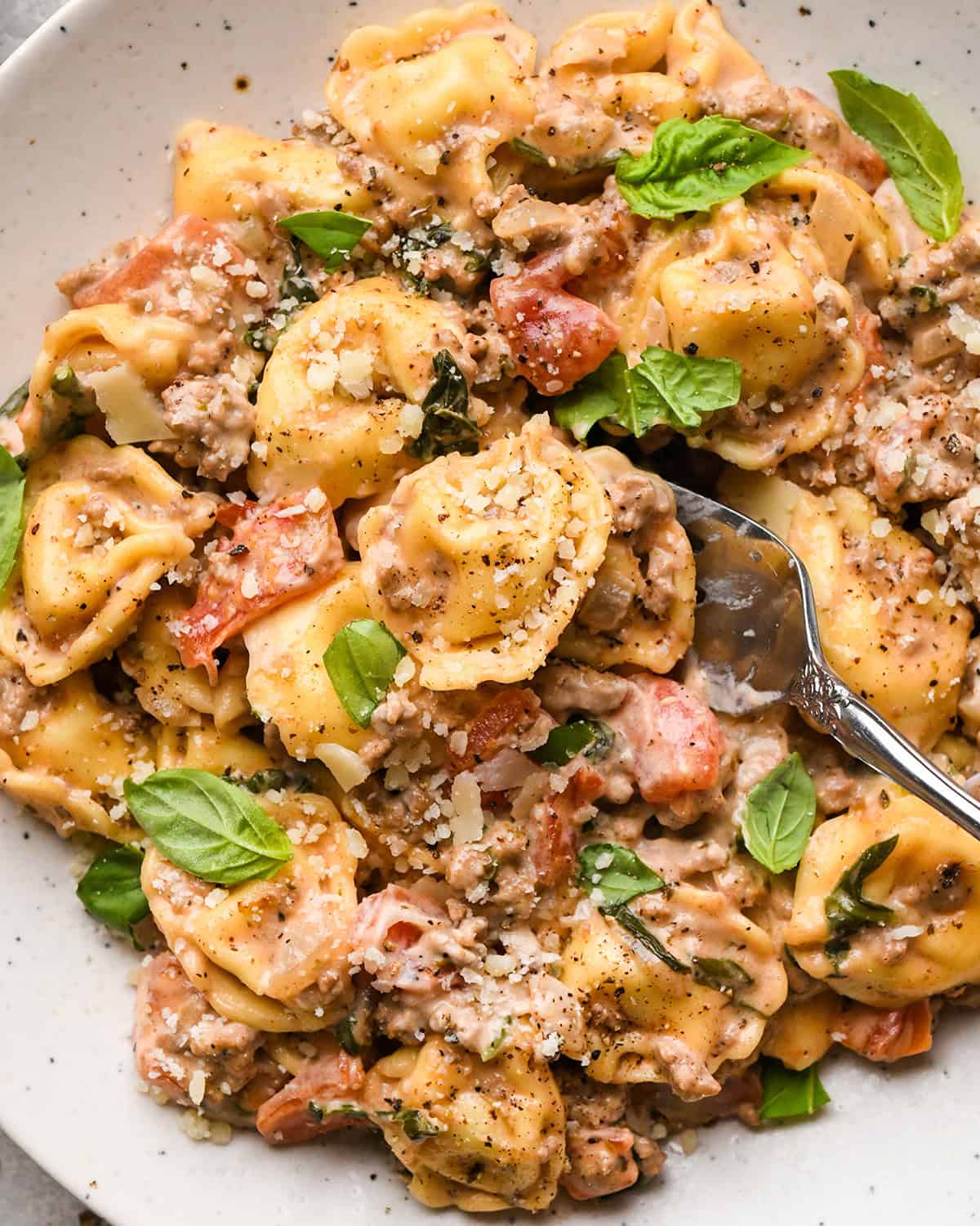 ground beef tortellini with diced tomatoes, fresh basil, and grated parmesan cheese,  in a white bowl with a silver fork. 