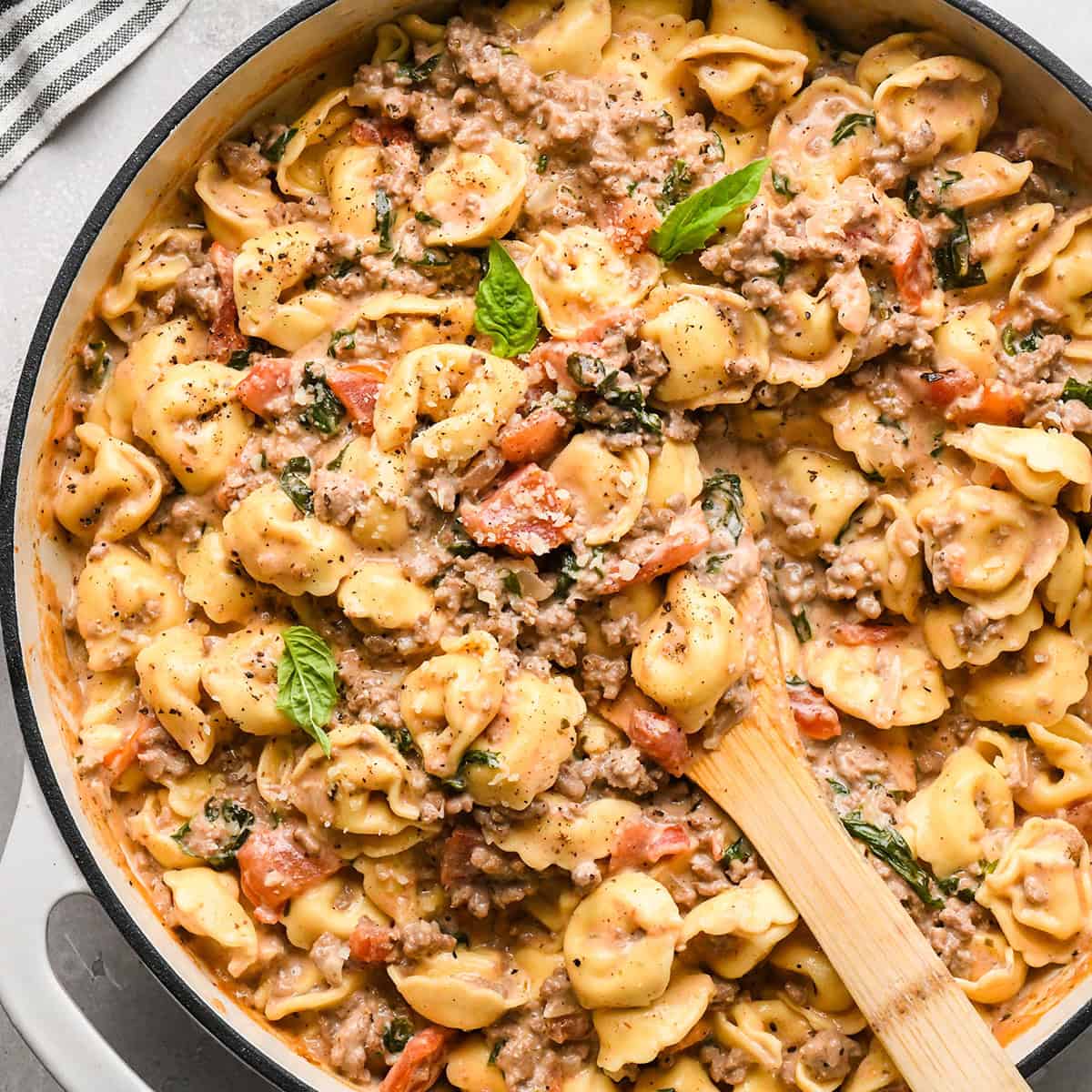 ground beef tortellini with diced tomatoes, fresh basil, and grated parmesan cheese in a skillet with a wooden spoon