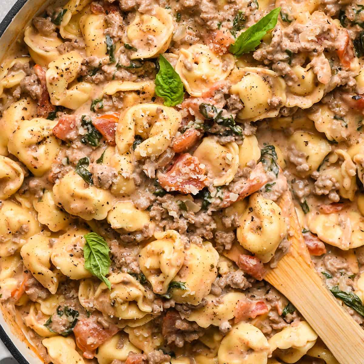ground beef tortellini with diced tomatoes, fresh basil, and grated parmesan cheese,  in a skillet with a wooden spoon