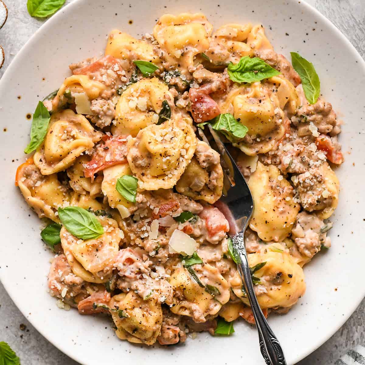 ground beef tortellini with diced tomatoes, fresh basil, and grated parmesan cheese,  in a white bowl with a silver fork. 