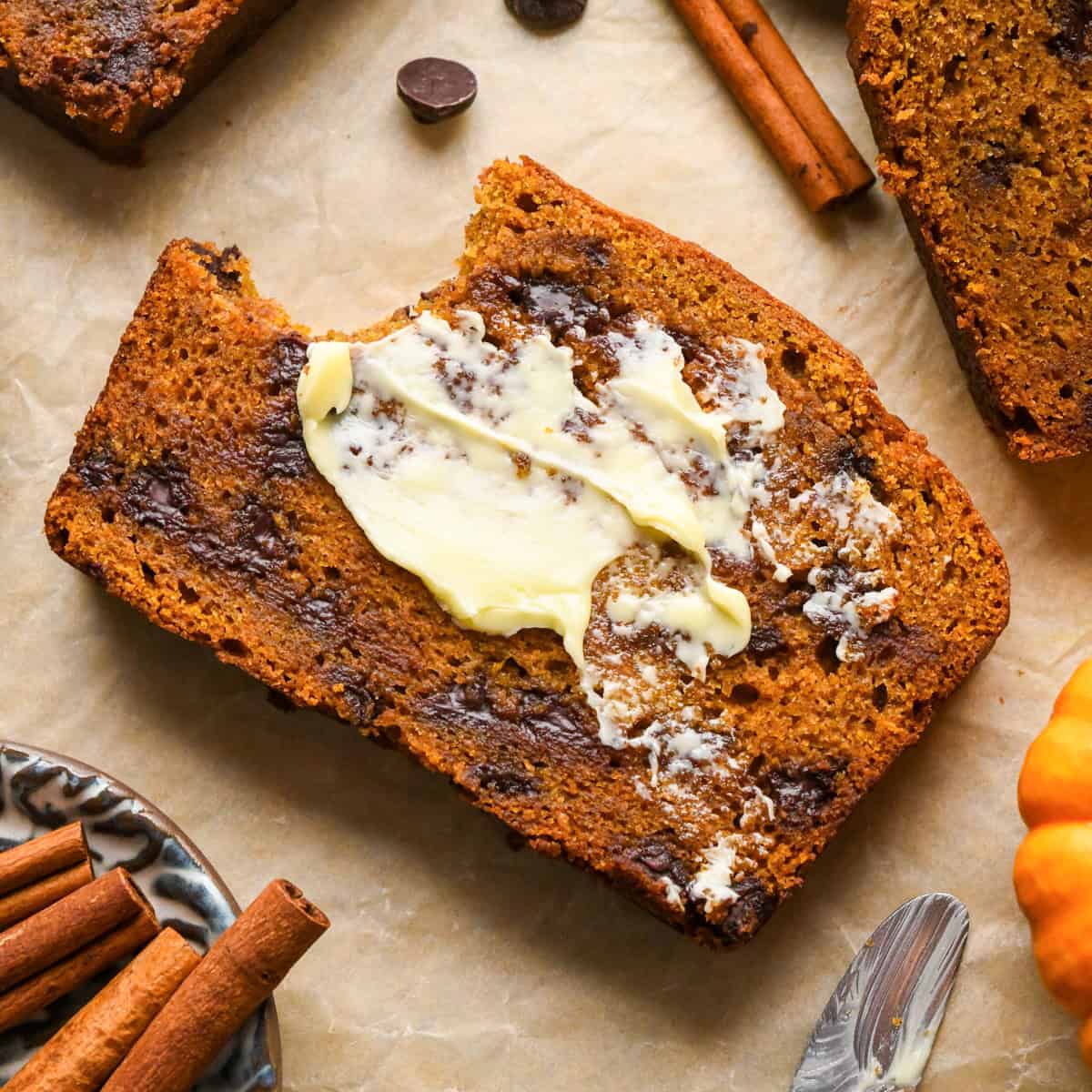 a slice of chocolate chip pumpkin bread with butter on it and a bite taken out of it on parchment paper, with melty chocolate chips surrounded by mini pumpkins, cinnamon sticks, and two more slices of bread