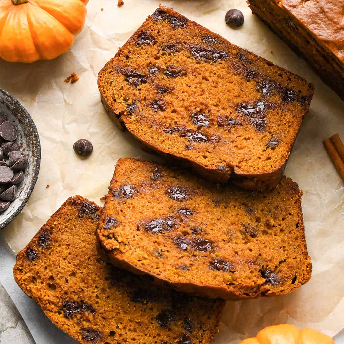 3 slices of chocolate chip pumpkin bread on parchment paper, with melty chocolate chips surrounded by mini pumpkins, cinnamon sticks, and a bowl of chocolate chips