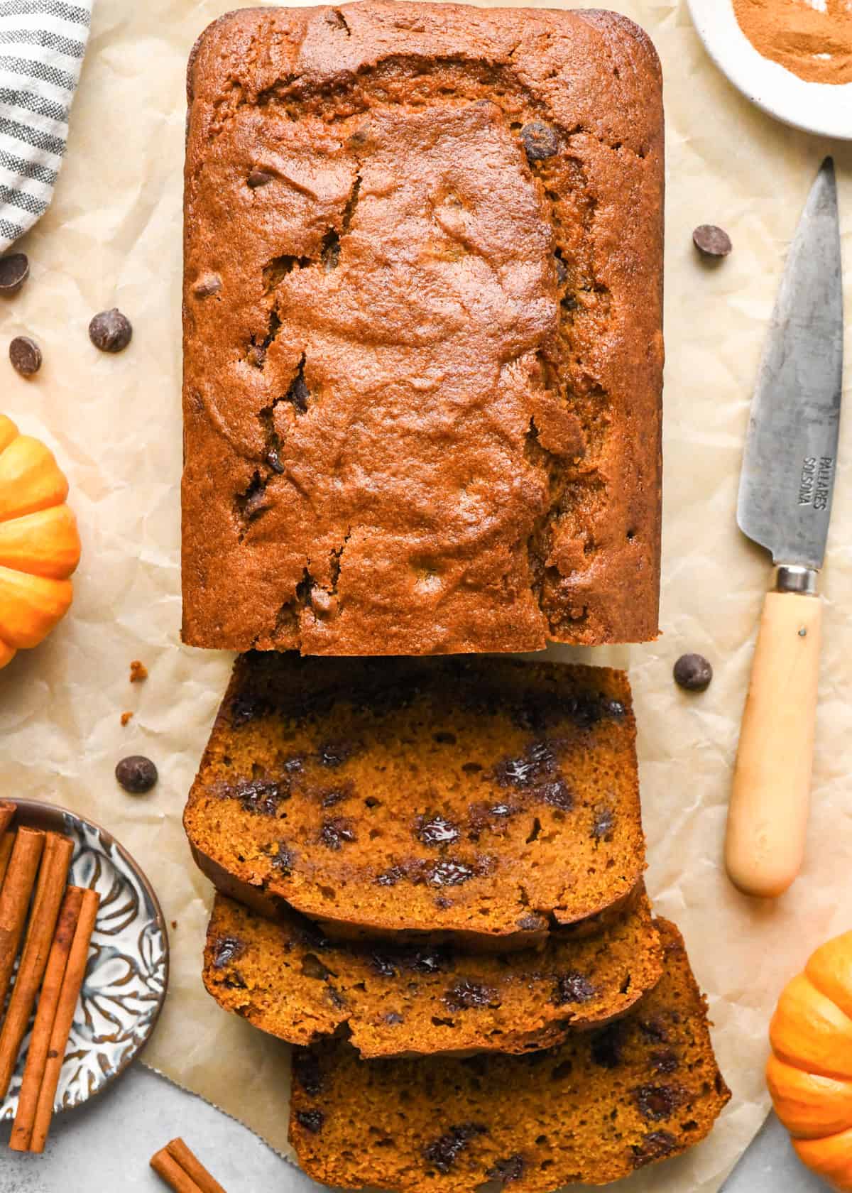 a loaf of chocolate chip pumpkin bread with 3 slices of cut out of it on parchment paper, with melty chocolate chips surrounded by mini pumpkins, cinnamon sticks, and a knife