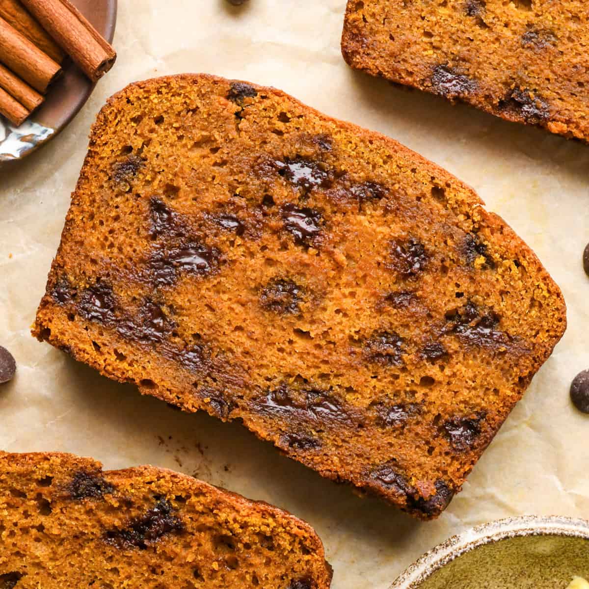 3 slices of Pumpkin Chocolate Chip Bread with melty chocolate chips on parchment paper surrounded by cinnamon sticks and chocolate chips